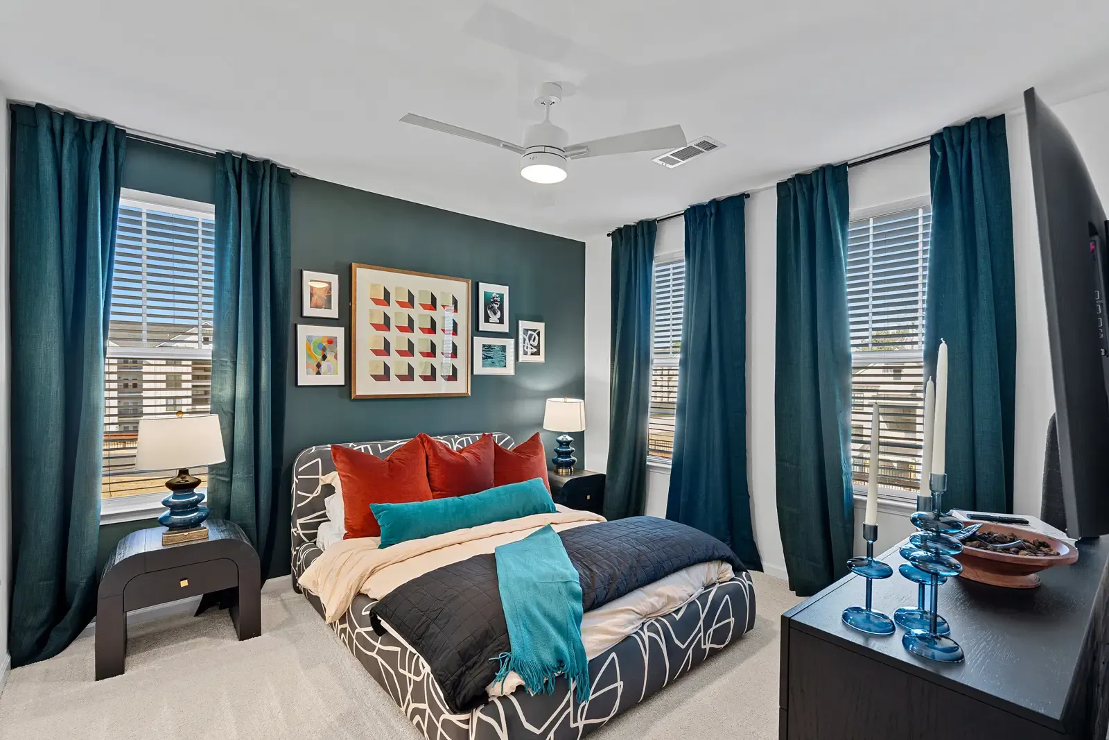Modern bedroom with dark teal curtains, a patterned bedspread, and red and teal pillows. Art frames adorn a dark accent wall, creating a cozy ambiance.