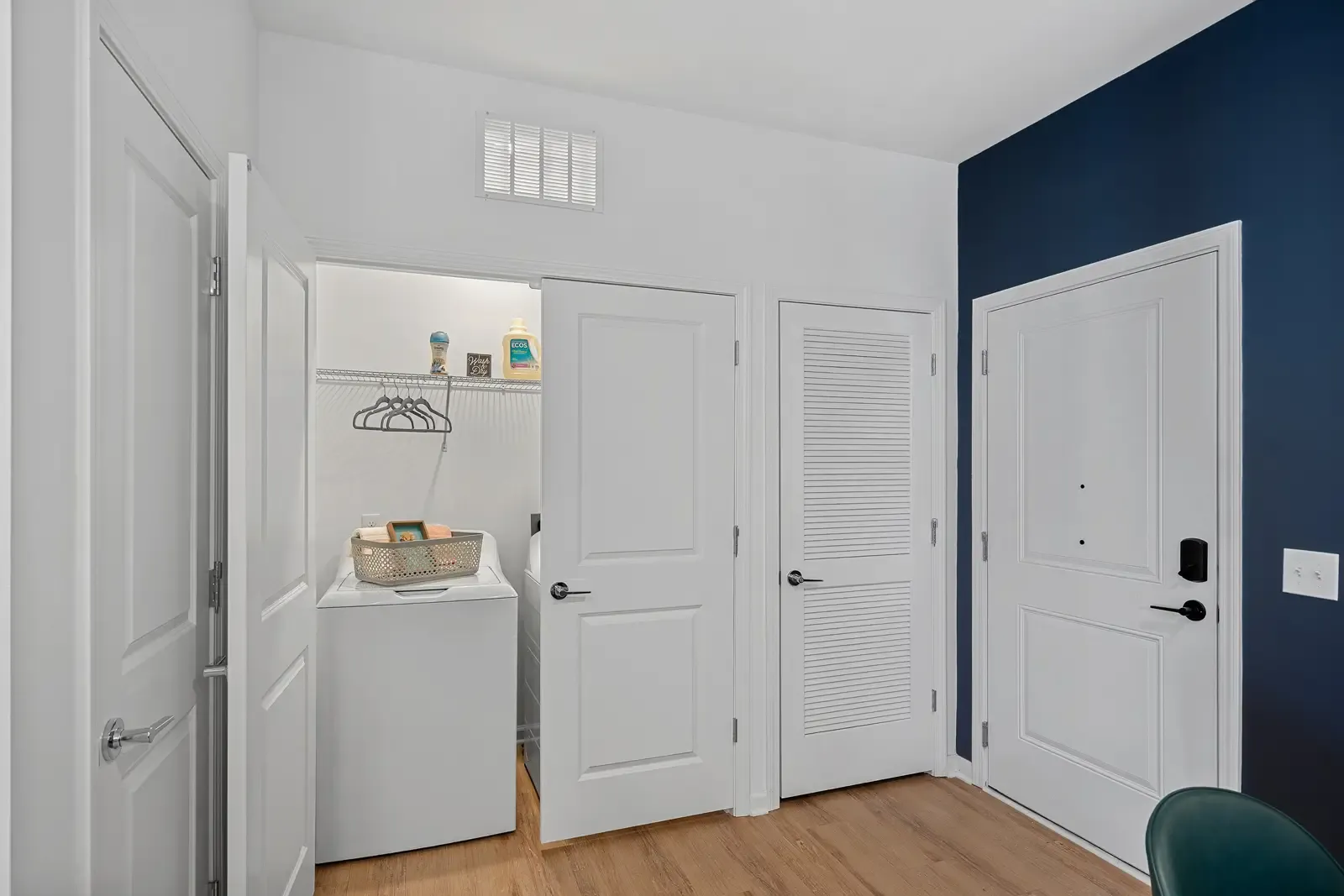 Laundry room with open door on left revealing a white washing machine, shelf with laundry supplies, hangers, and basket. Simple, organized, and clean.