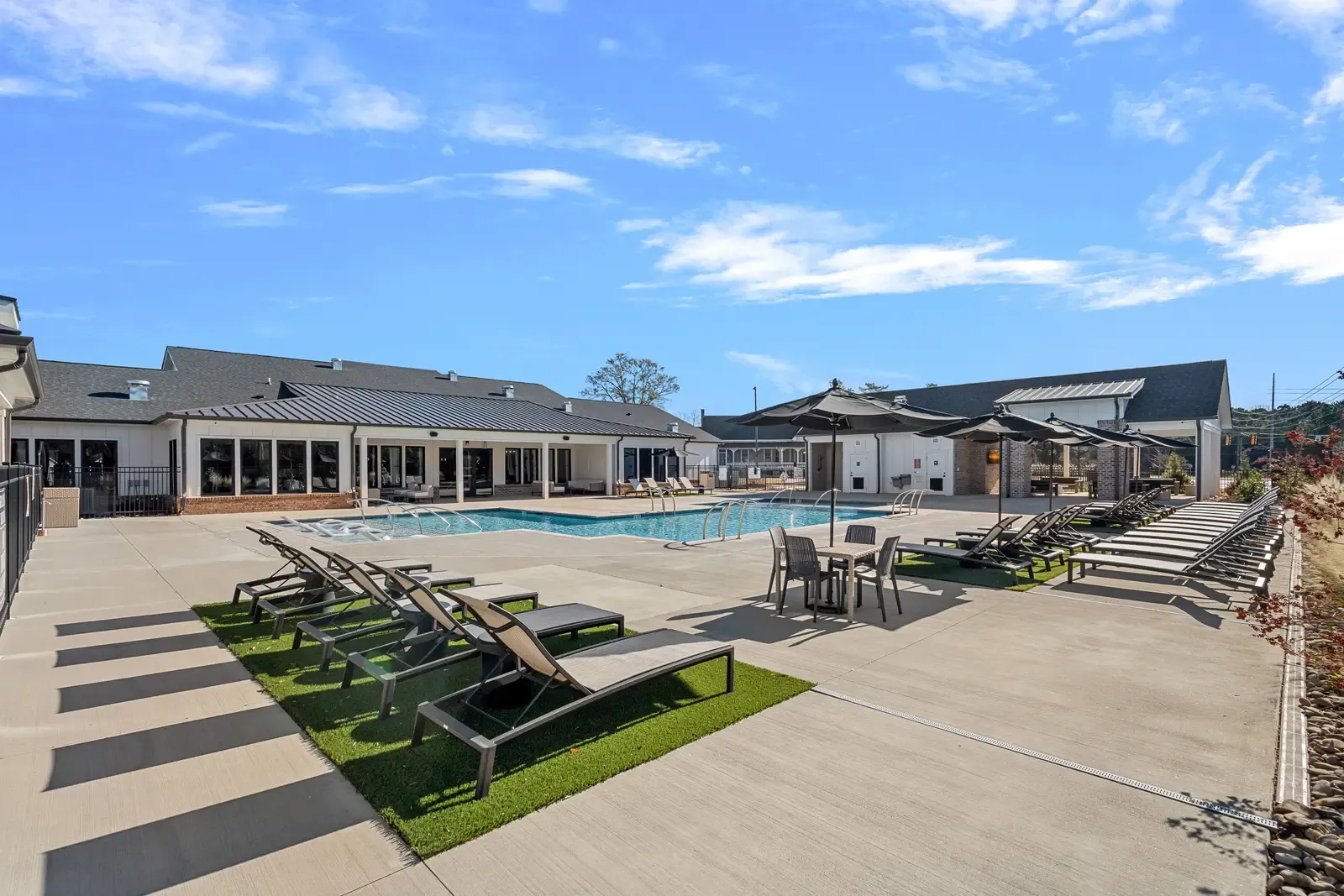 Outdoor pool area with modern sun loungers and umbrellas on a sunny day. The serene setting is surrounded by stylish buildings under a clear blue sky.