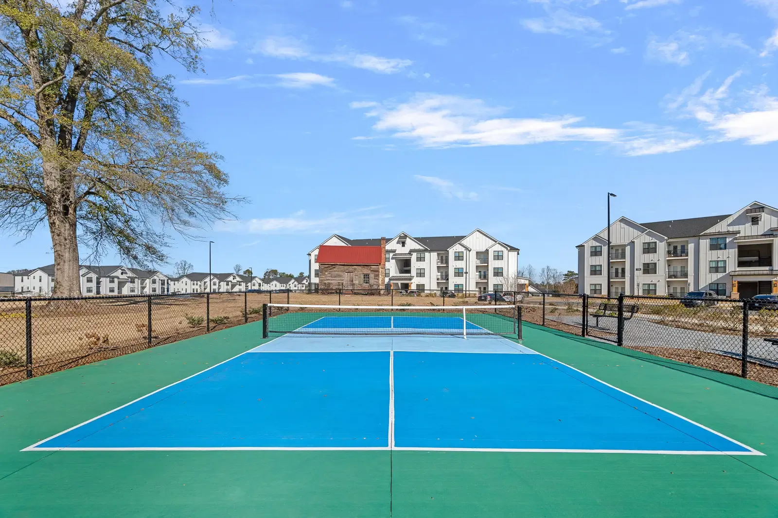 A pickleball court with bright blue surfaces and green borders is in the foreground, surrounded by a black fence. Modern apartments stand under a clear blue sky.