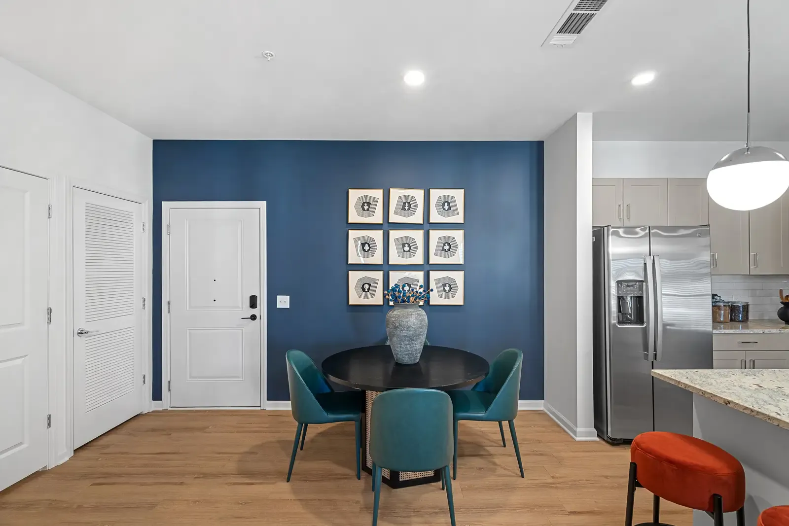 Modern dining area featuring a round black table with teal chairs against a blue accent wall. Nine minimalist art pieces and a vase add elegance.