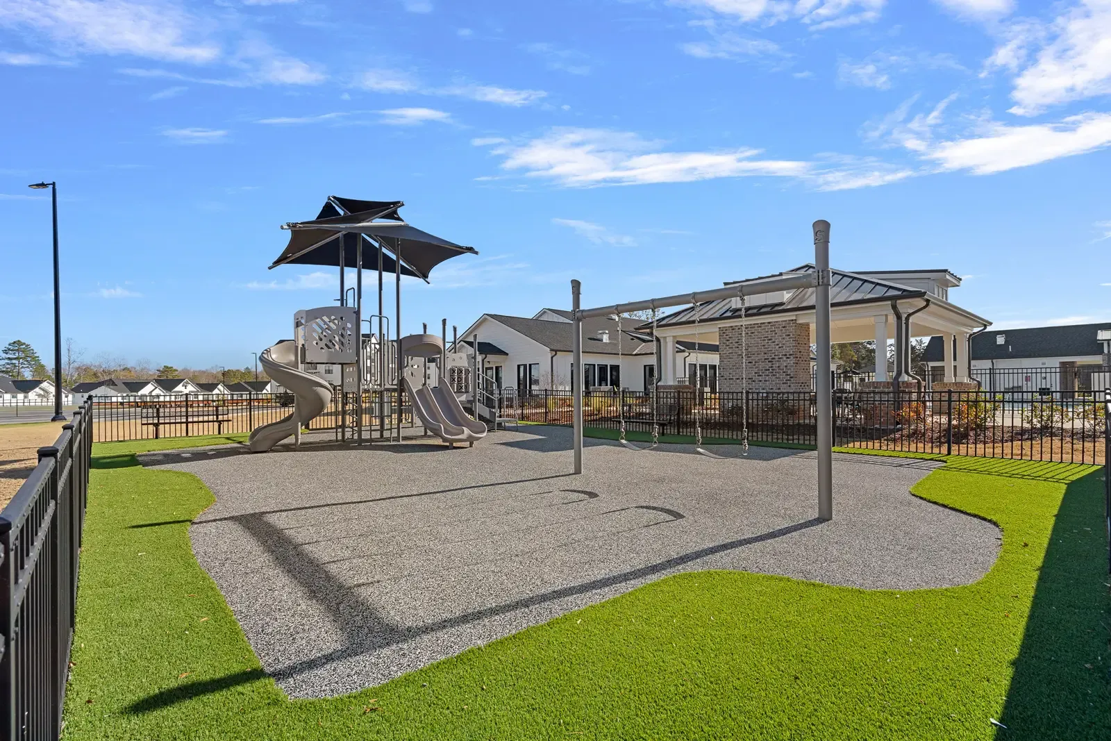 A sunny playground features a climbing structure with slides, surrounded by green artificial turf. Nearby houses and blue sky create a welcoming feel.