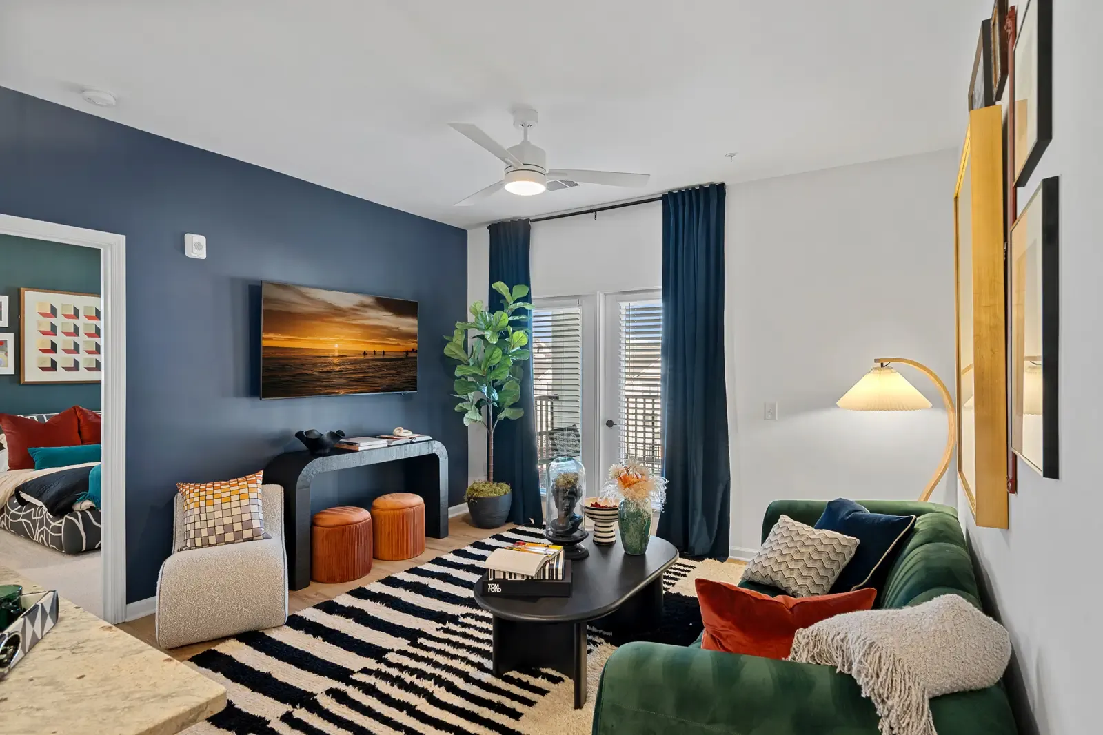 Modern living room with dark blue accent wall, wall-mounted TV, and striped rug. Green sofa with colorful pillows adds warmth. Glass doors let in light.