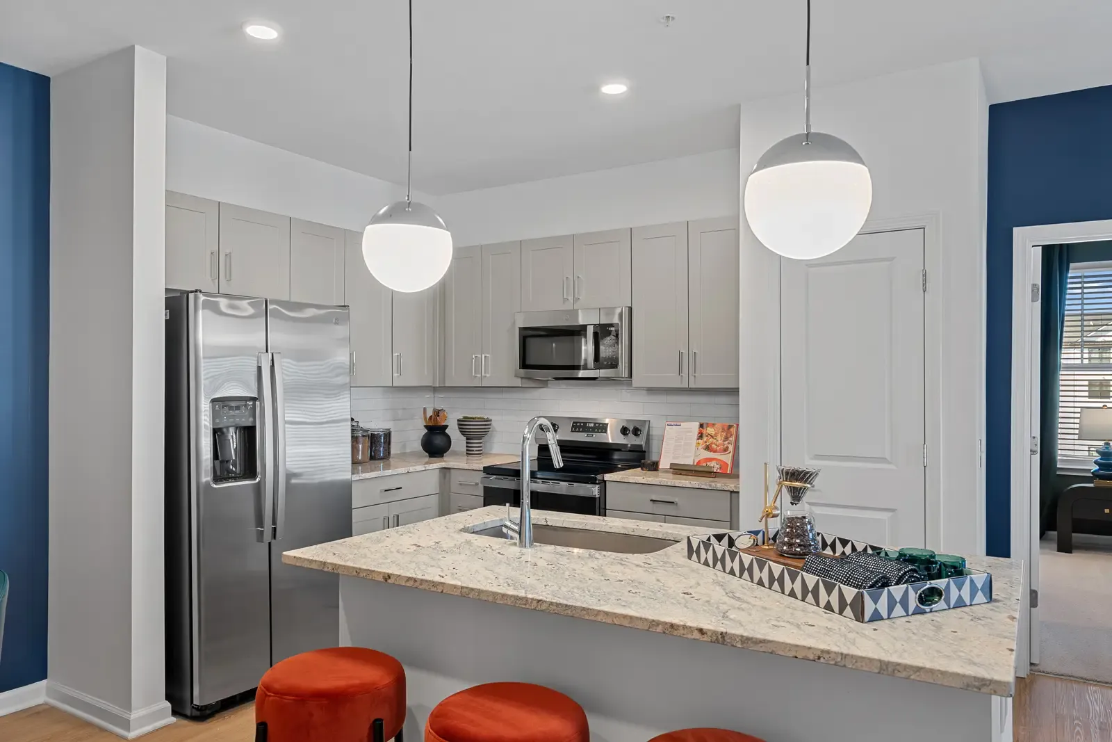 Modern kitchen with stainless steel appliances, granite countertops, stylish orange stools, and pendant lighting against a blue accent wall.