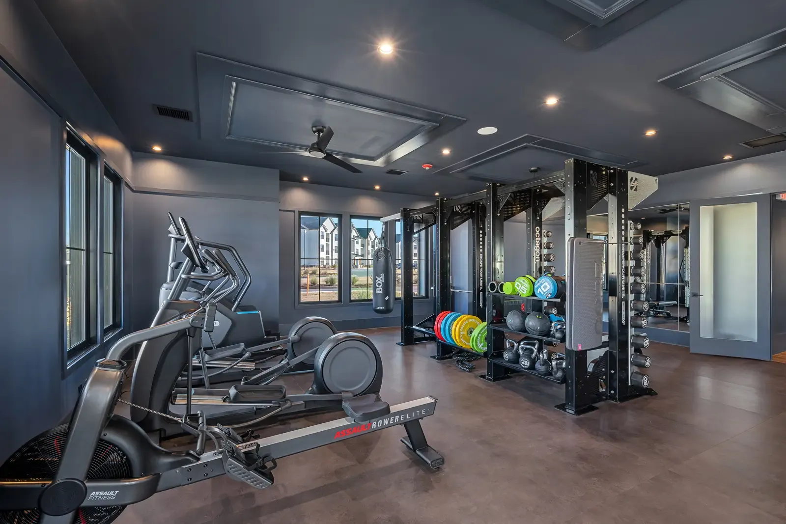 A modern gym with dark walls, multiple exercise machines including bikes and rowing machines to the left, and colorful weights and kettlebells on racks to the right.